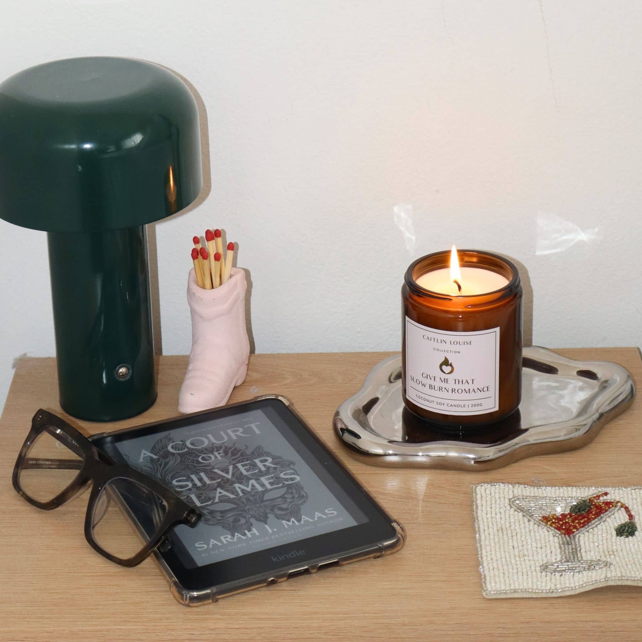 Tabletop scene with a lit candle, book, glasses, and small lamp.