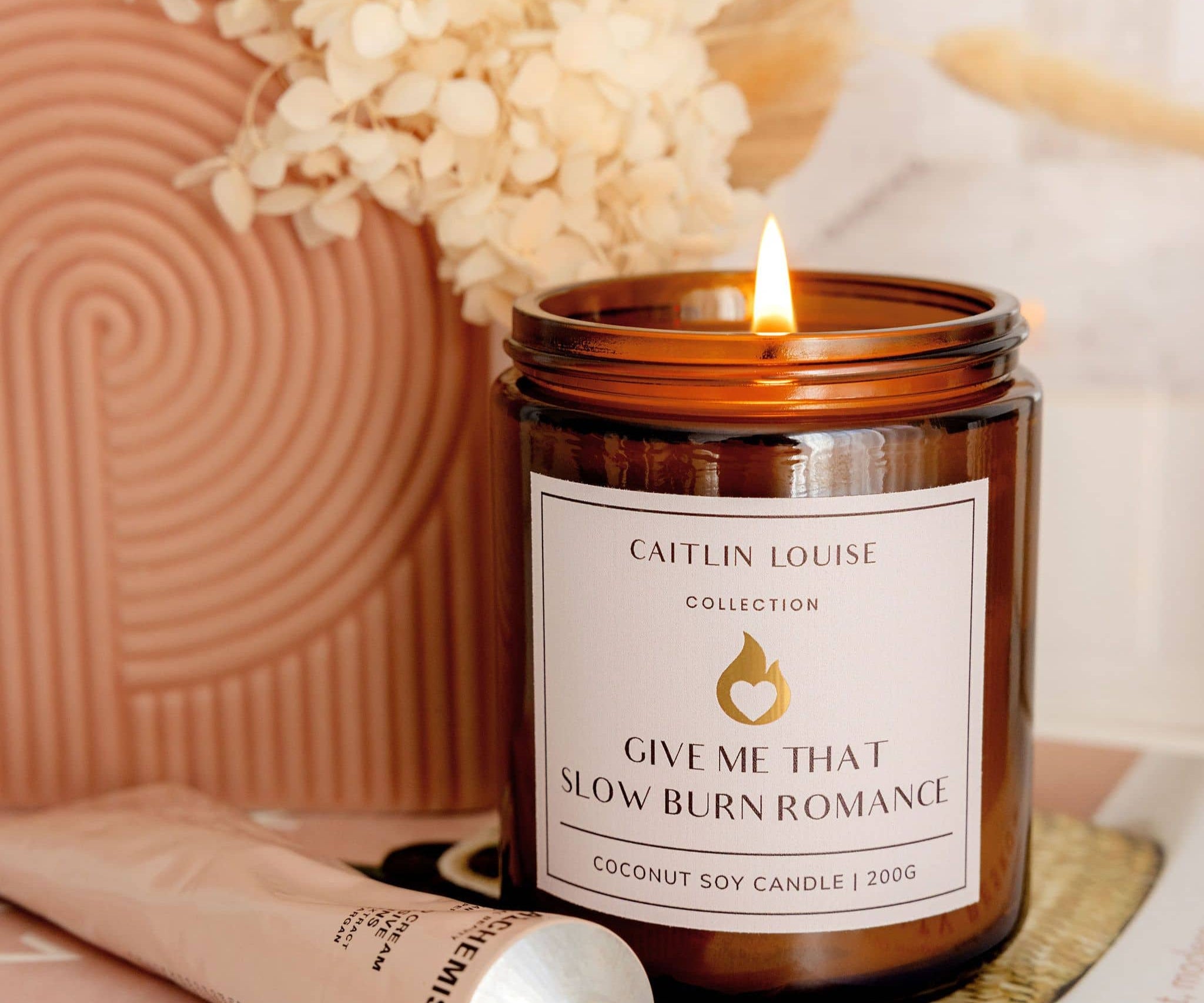 Scented Candle with a label 'Give Me That Slow Burn Romance' from Caitlin Louise Collection, surrounded by decorative elements.