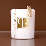 Caitlin Louise Collection candle with gold logo and gold jewelry on a brown background