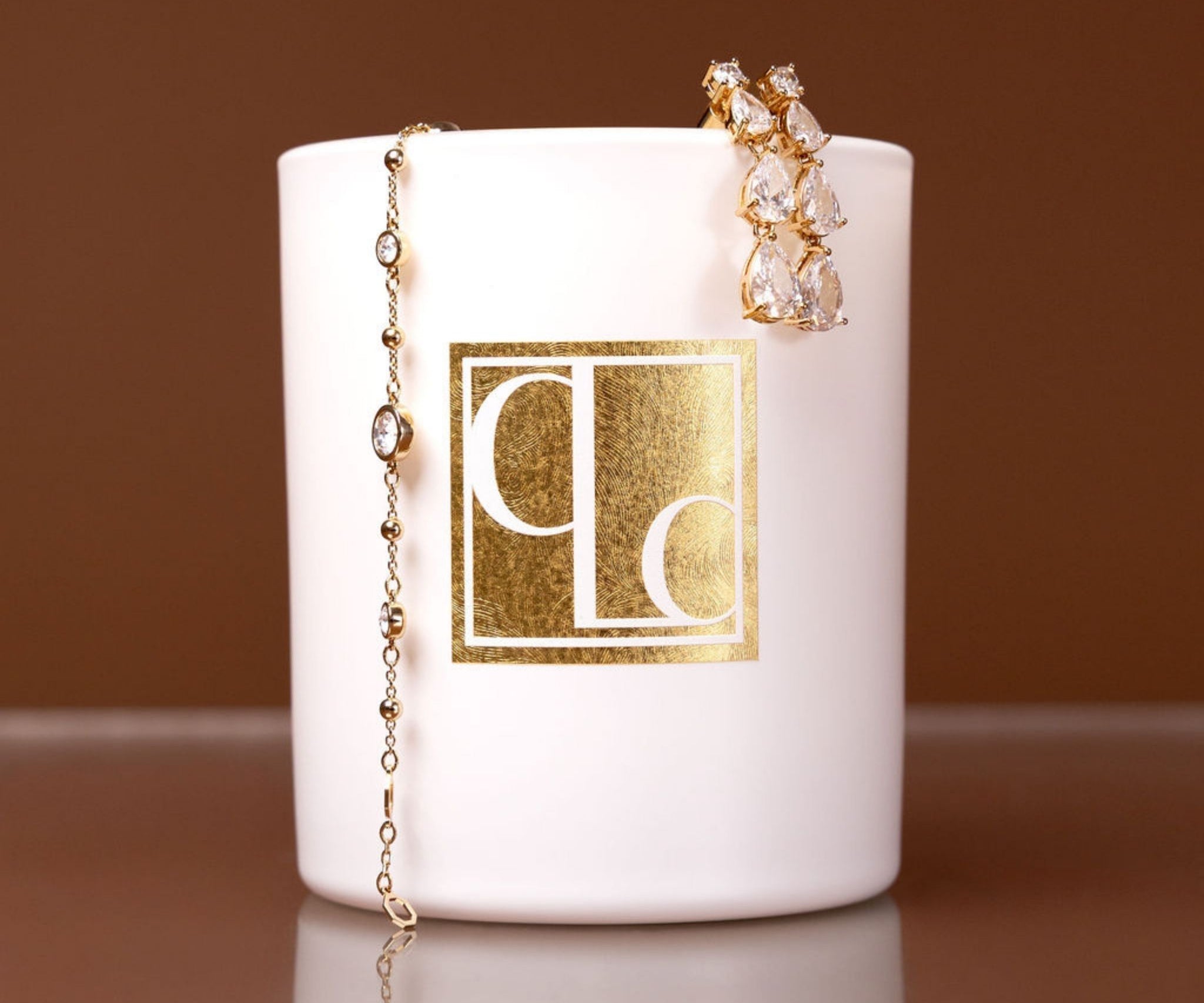 Caitlin Louise Collection candle with gold logo and gold jewelry on a brown background