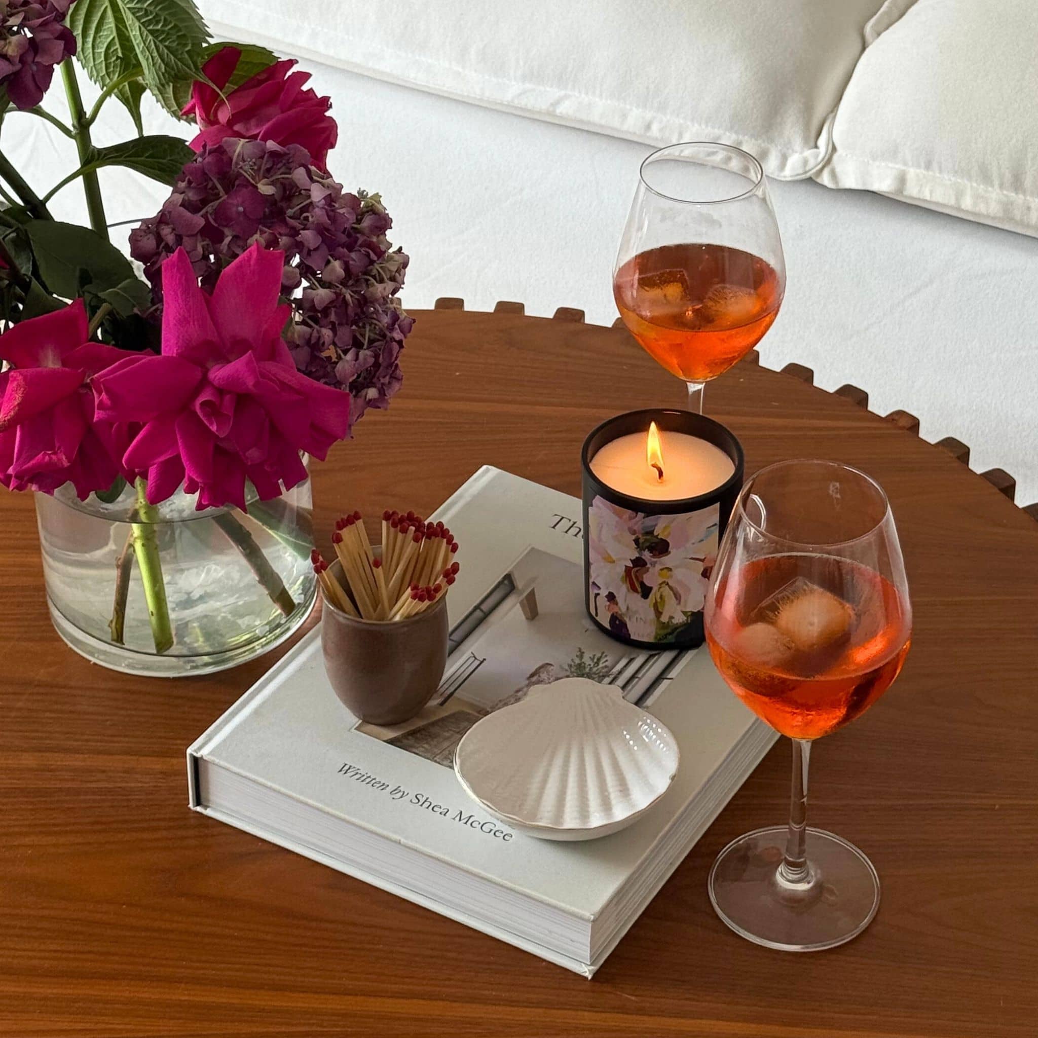 Two glasses of spritz, a lit candle, a book, a small bowl, and a vase with pink flowers on a wooden table.