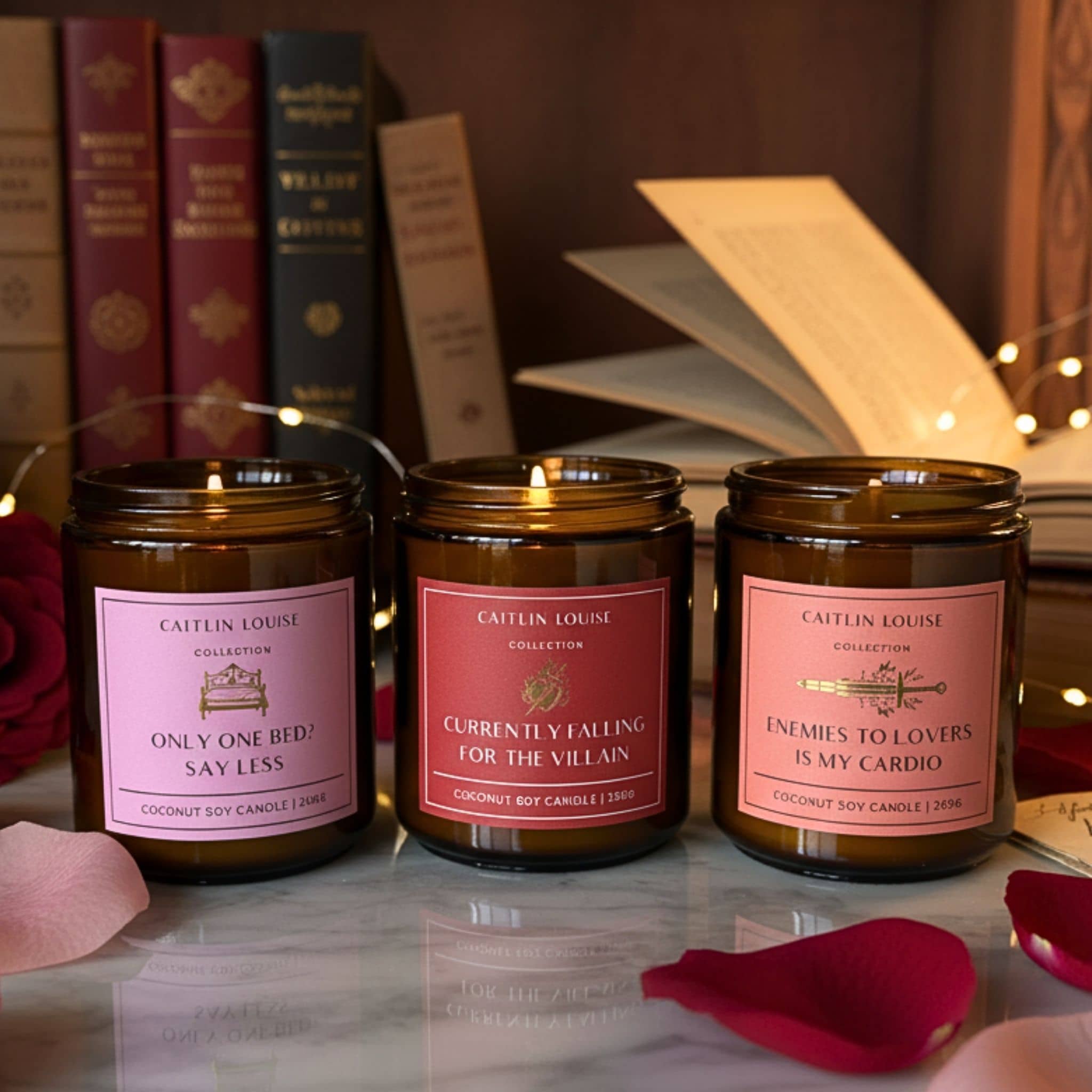 Three candles with pink labels on a marble surface with books and rose petals in the background.