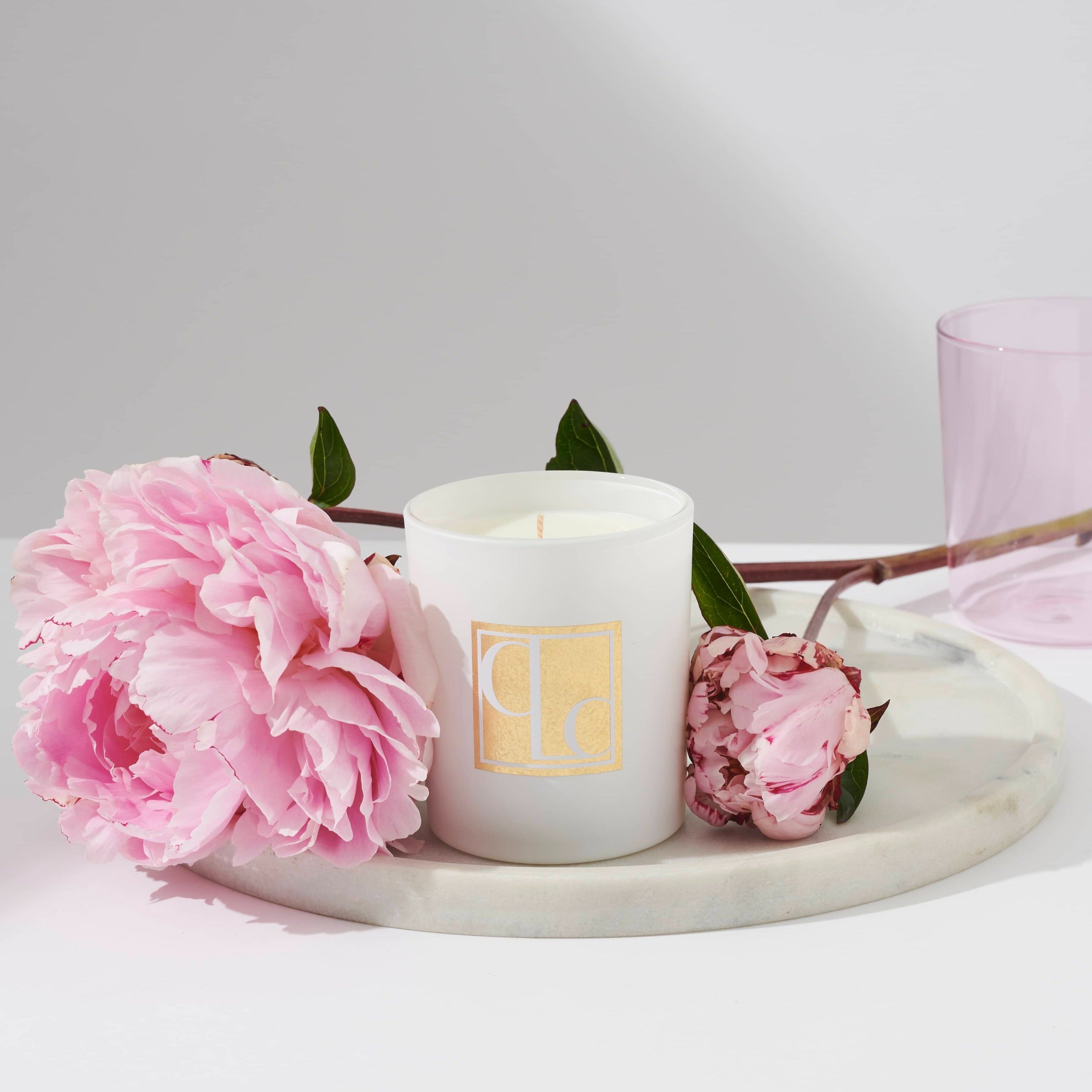 A white candle with the Lychee Peony scent, placed on a marble surface, surrounded by pink peony flowers and a pink glass.