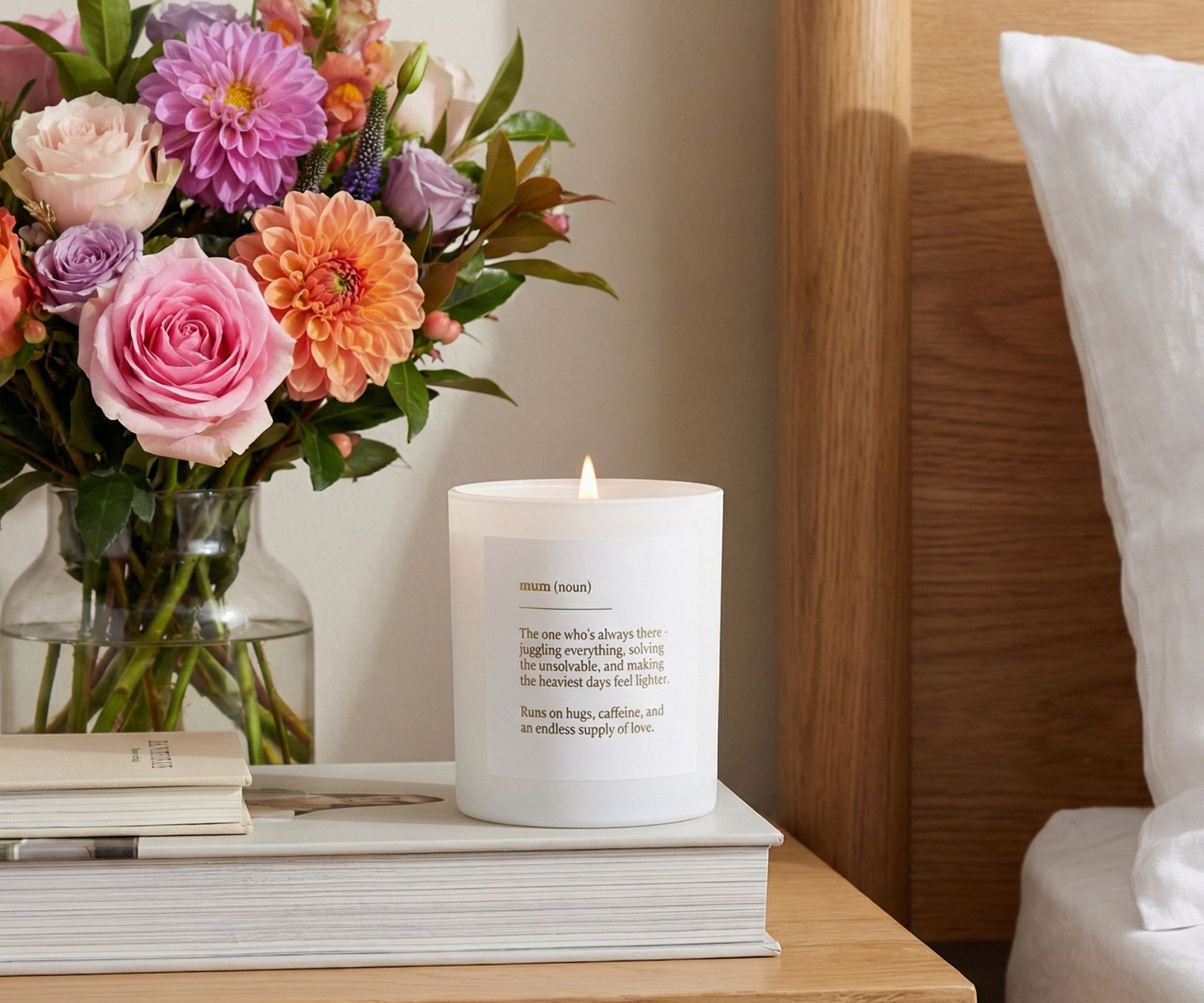 Candle with a message on a stack of books next to a vase of flowers on a wooden nightstand.