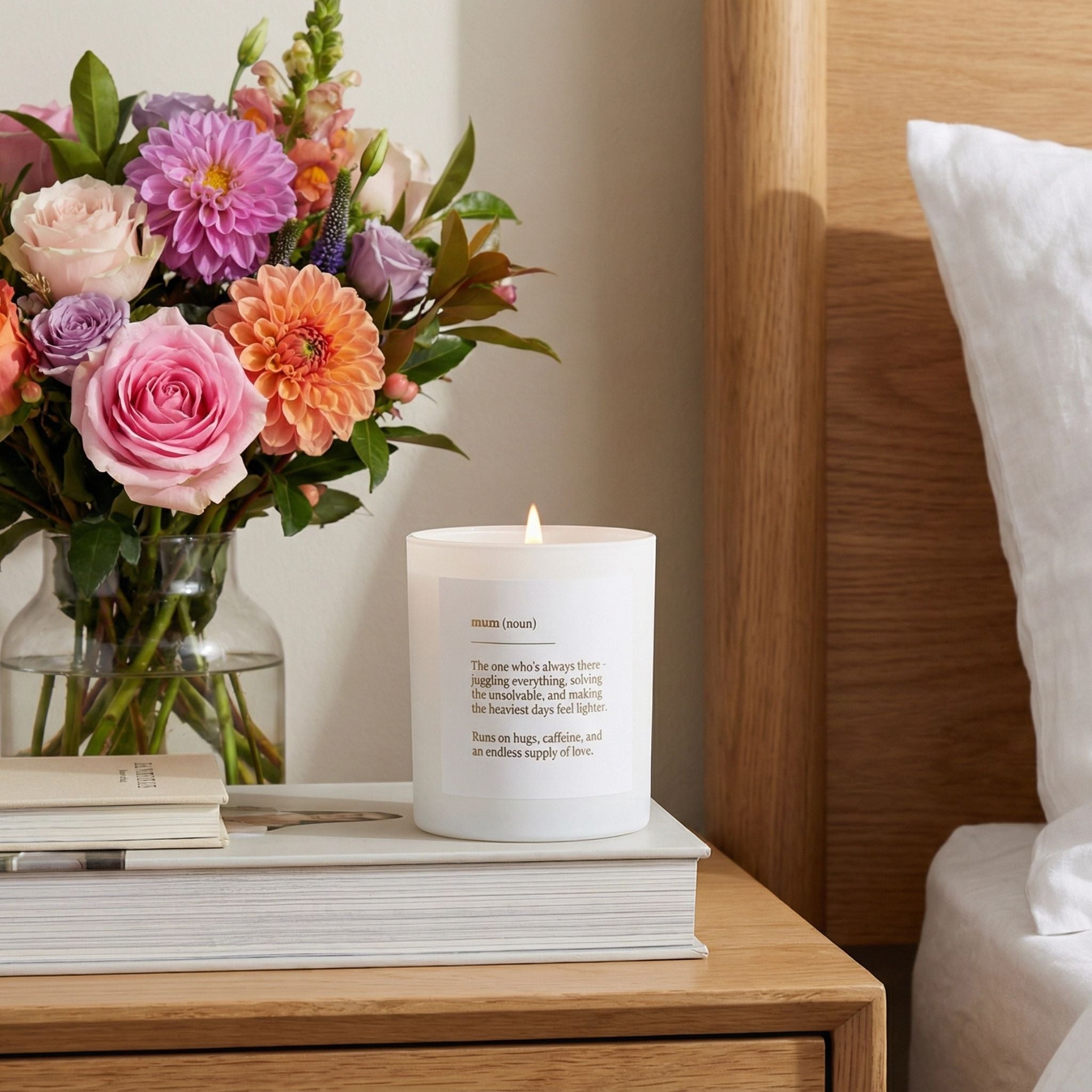 Candle with a message on a stack of books next to a vase of flowers on a wooden nightstand.
