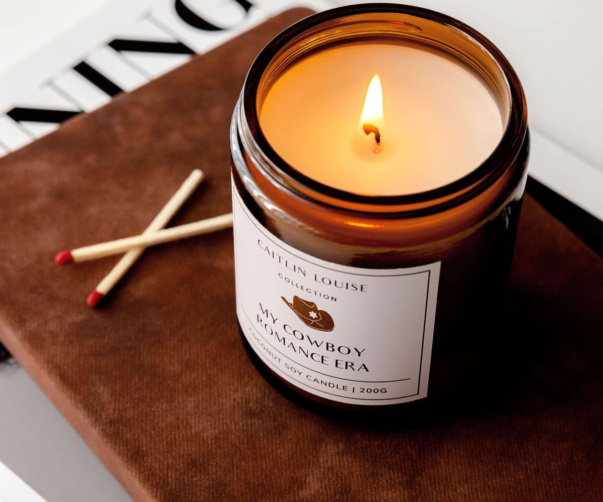 Candle in a glass jar labeled 'My Cowboy Romance Era' on a leather surface with matches.