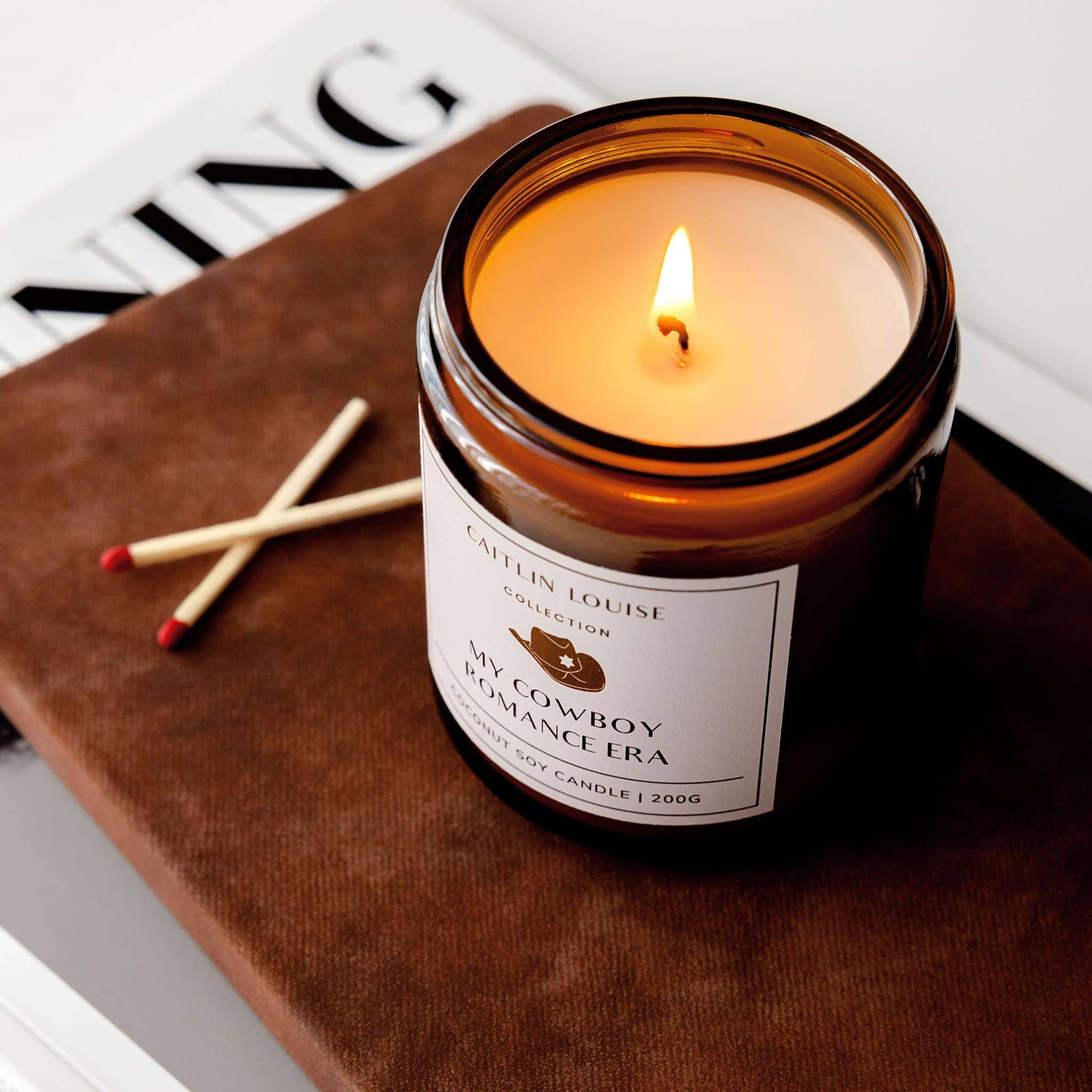 Candle in a glass jar labeled 'My Cowboy Romance Era' on a leather surface with matches.