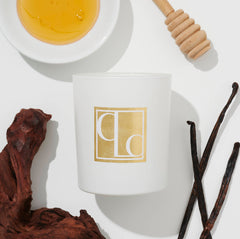 A white candle with the brand mark 'CLC' in gold, surrounded by honey, a honey dipper, dried fruits, and vanilla pods on a light background.