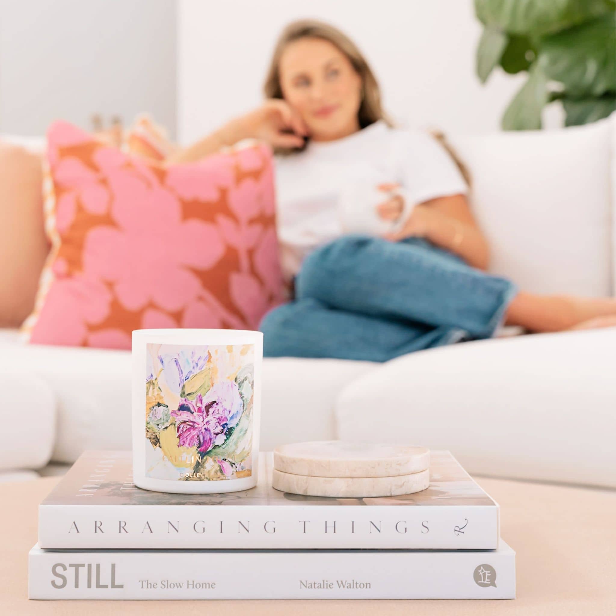 Caitlin Louise Collection's Patchouli and Sandalwood candle sitting on stack of books with woman on the lounge in the background.
