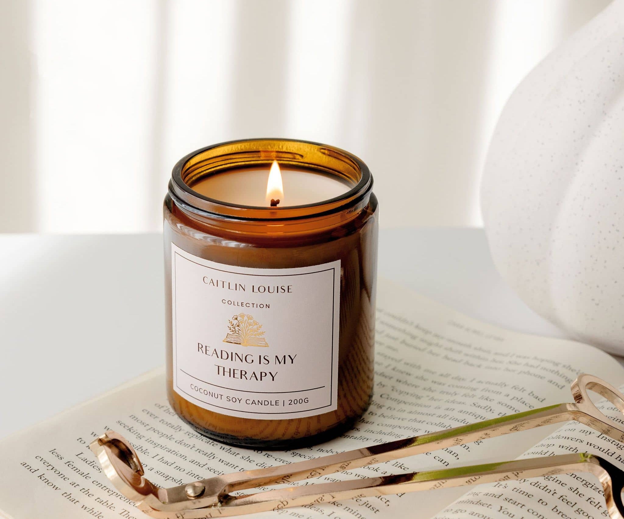 Candle with 'Reading is my Therapy' label on an open book, surrounded by wick trimmers and a white vase.