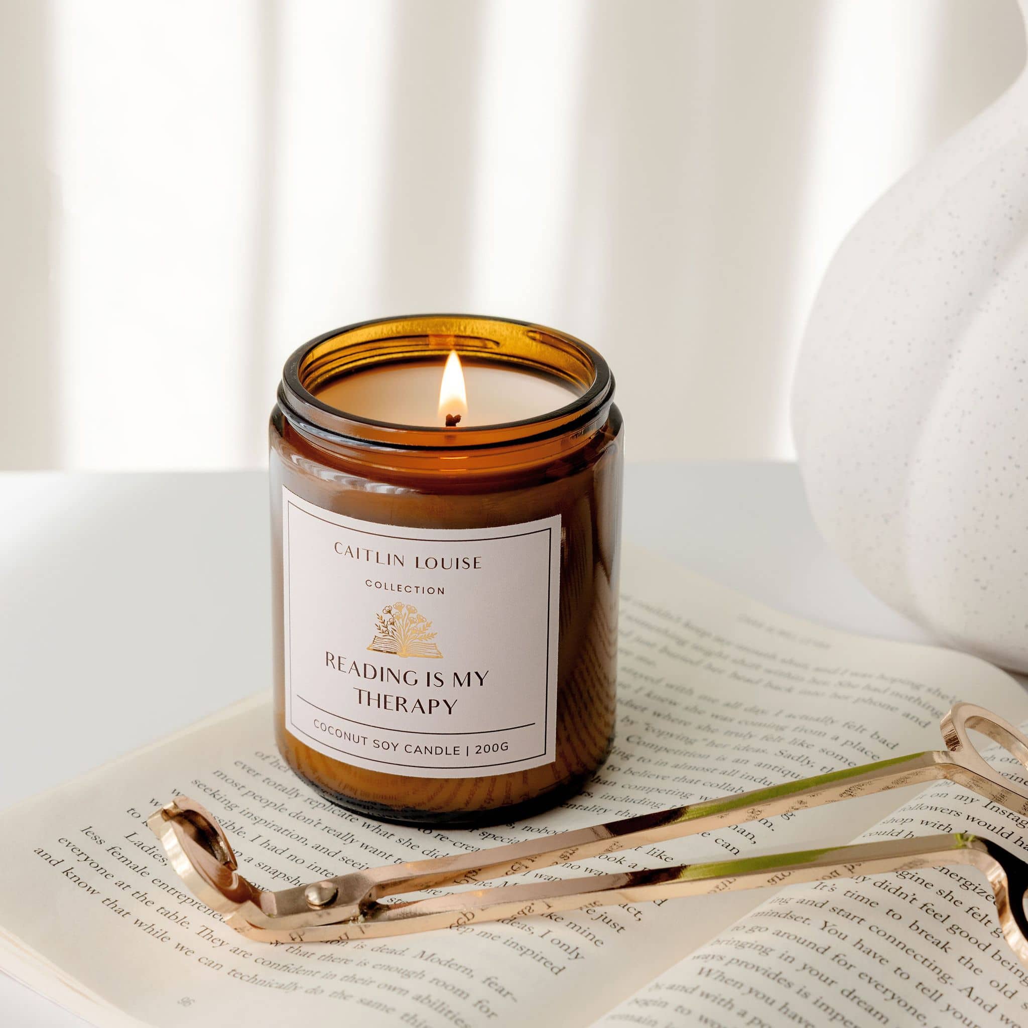 Candle with 'Reading is my Therapy' label on an open book, surrounded by wick trimmers and a white vase.