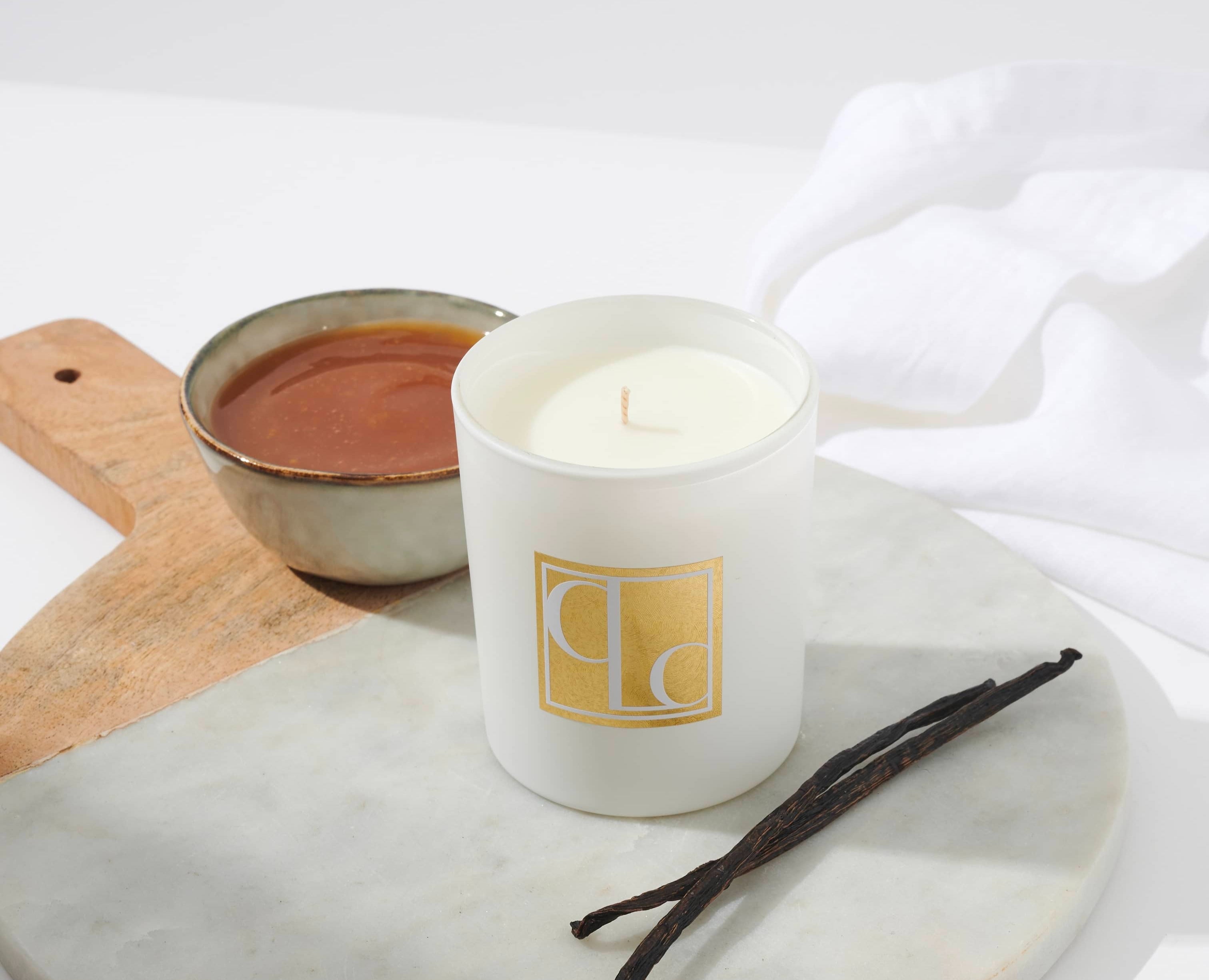 Caitlin Louise Collection Vanilla Caramel Scented Candle on board with bowl of caramel in background and vanilla sticks in the foreground.