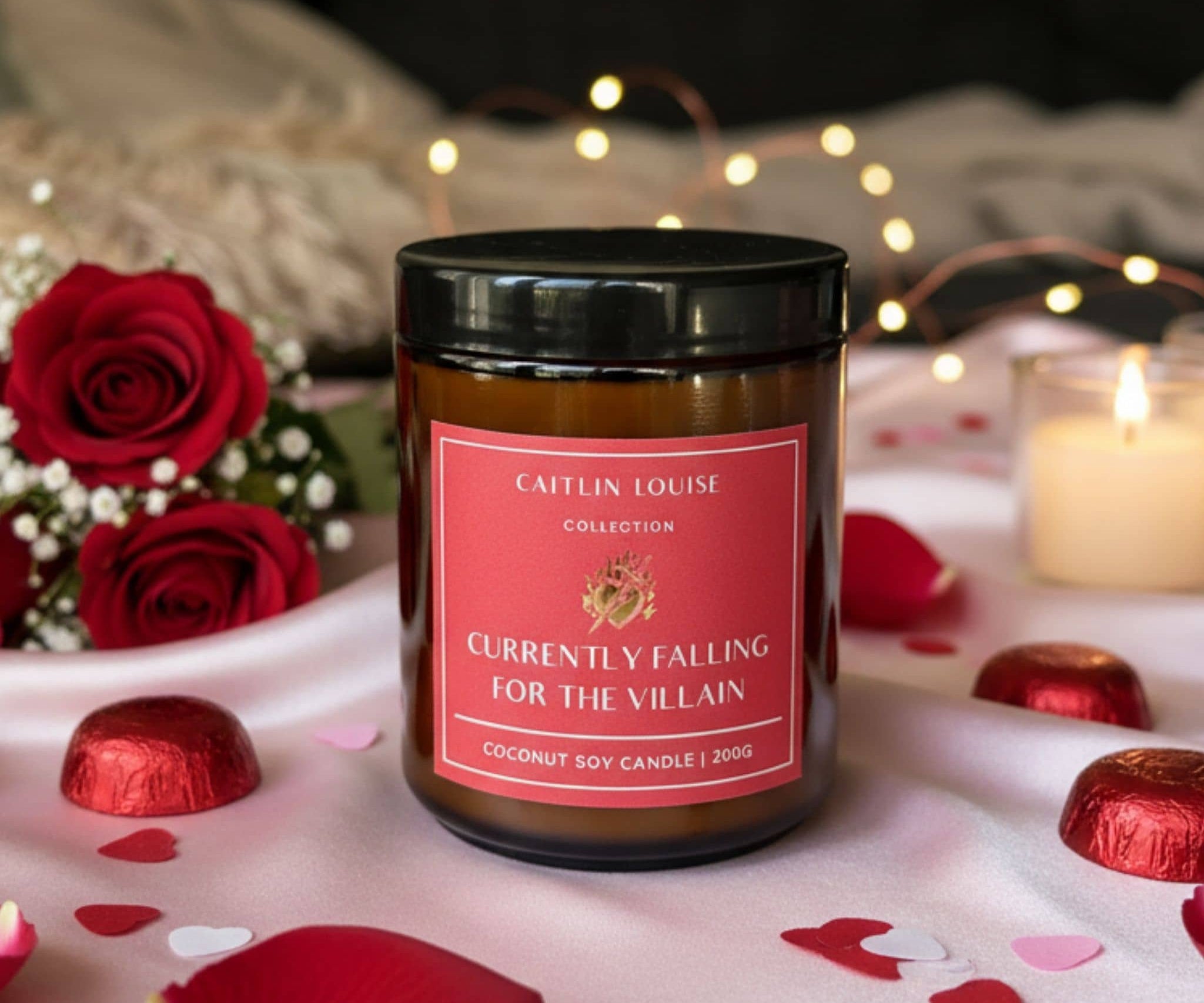 Candle with a red label on a romantic setting with roses and chocolate, featuring 'Caftin Louise Collection'.