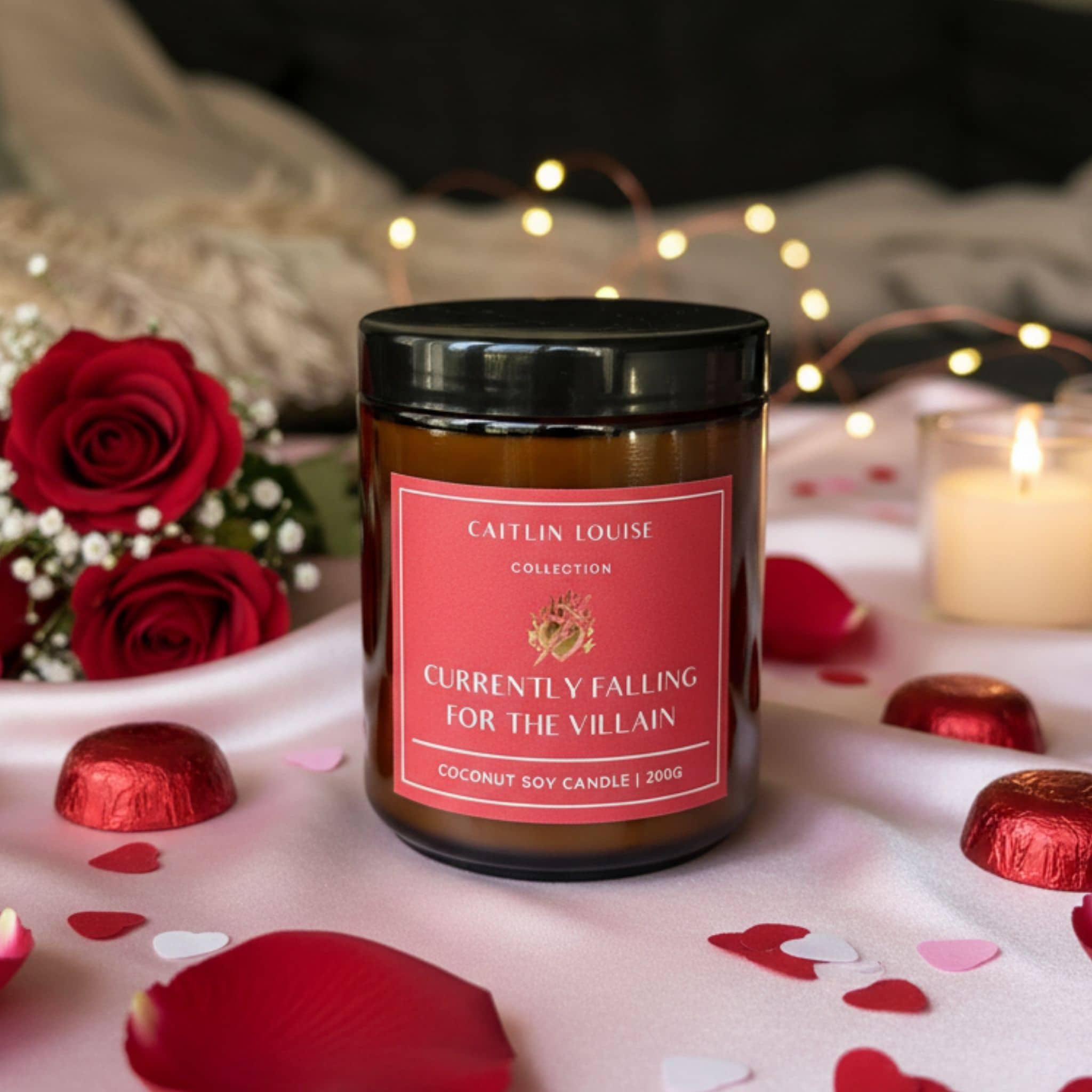 Candle with a red label on a romantic setting with roses and chocolate, featuring 'Caftin Louise Collection'.