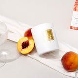 White candle with gold logo, sliced peach, and whole peach on a white surface