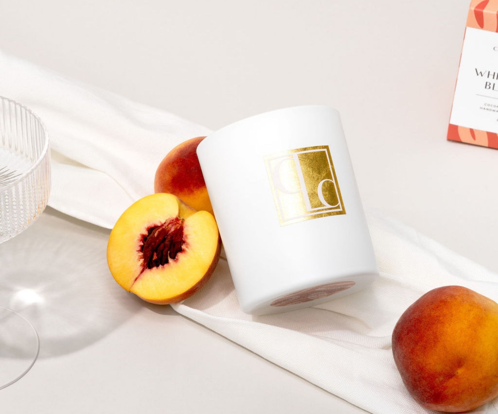 White candle with gold logo, sliced peach, and whole peach on a white surface