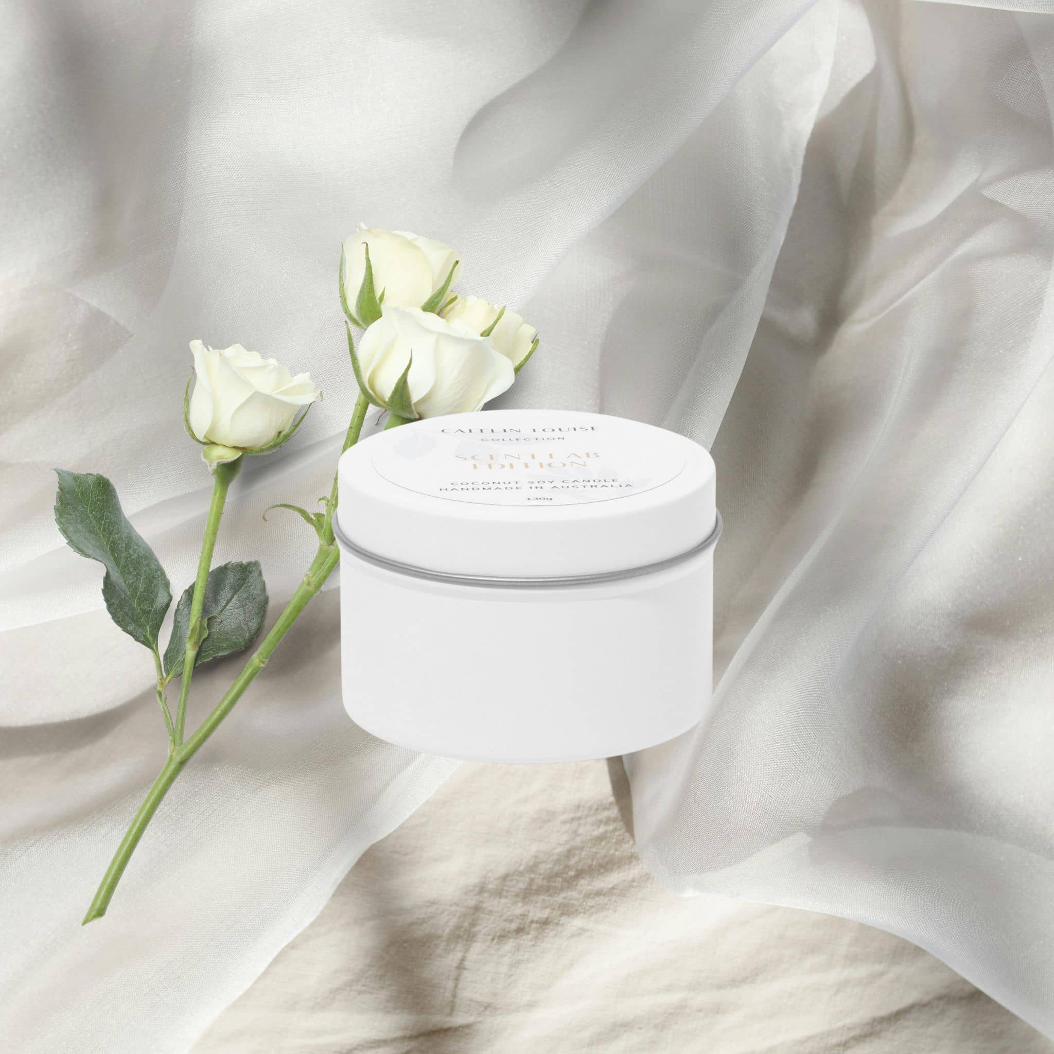 White travel tin candle with a lid on a white fabric background with white roses.